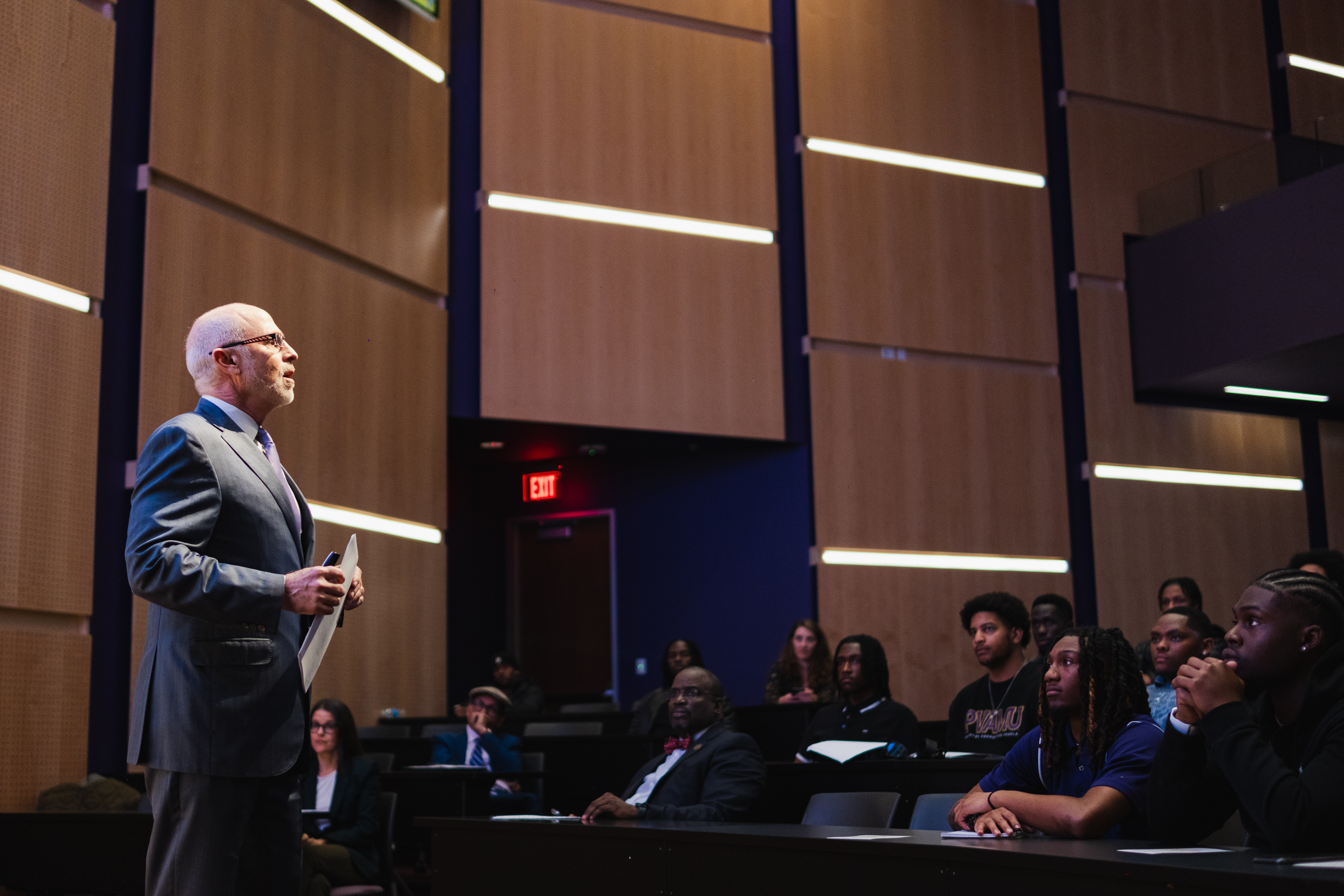 Faculty Education Programs Male professor in suit lecturing to attentive students in a modern university classroom with wood panel walls and overhead lighting.