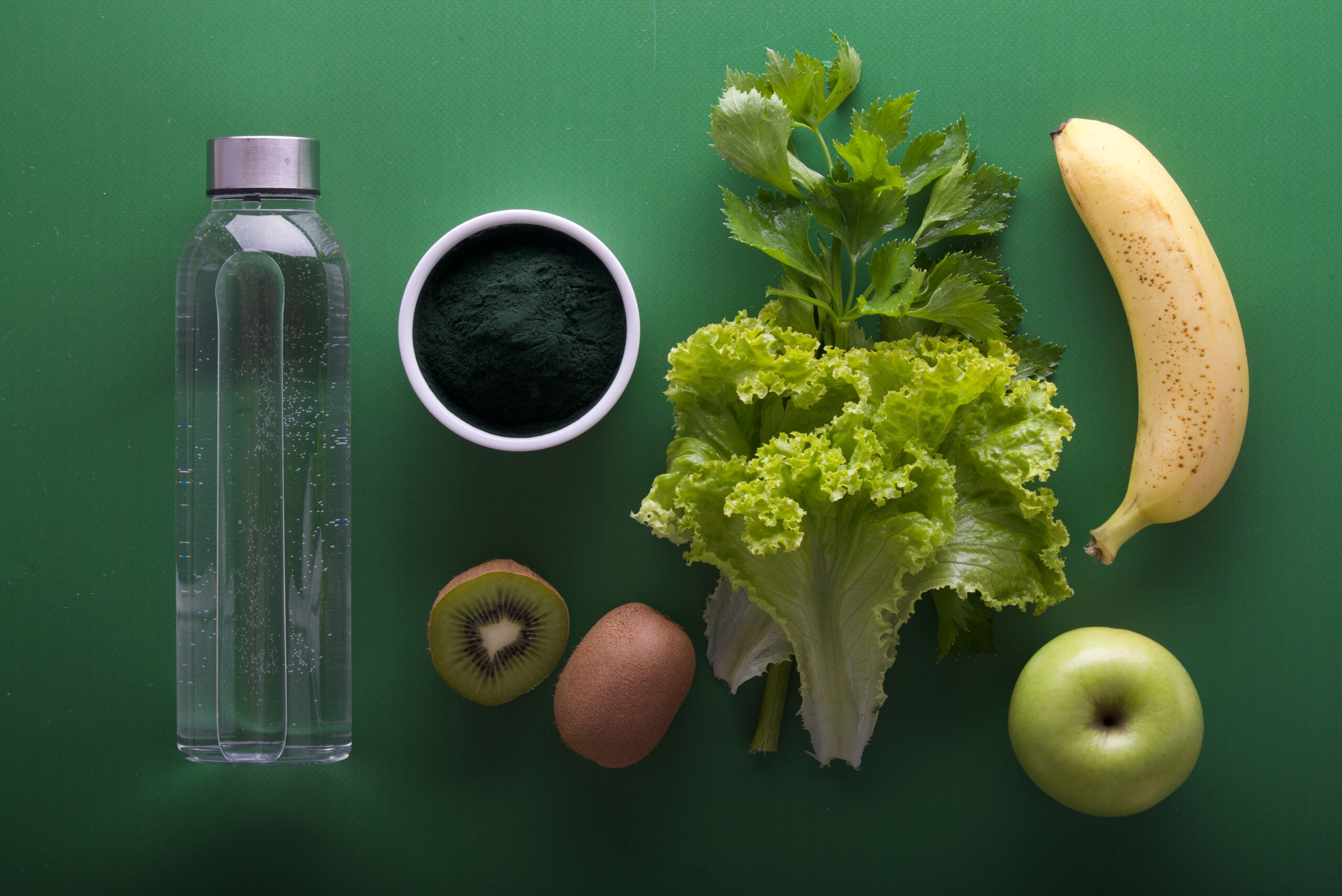 A bottle of water, a sliced kiwi, a wedge of Romaine lettuce, a granny smith apple, and a banana lined up on a green background.