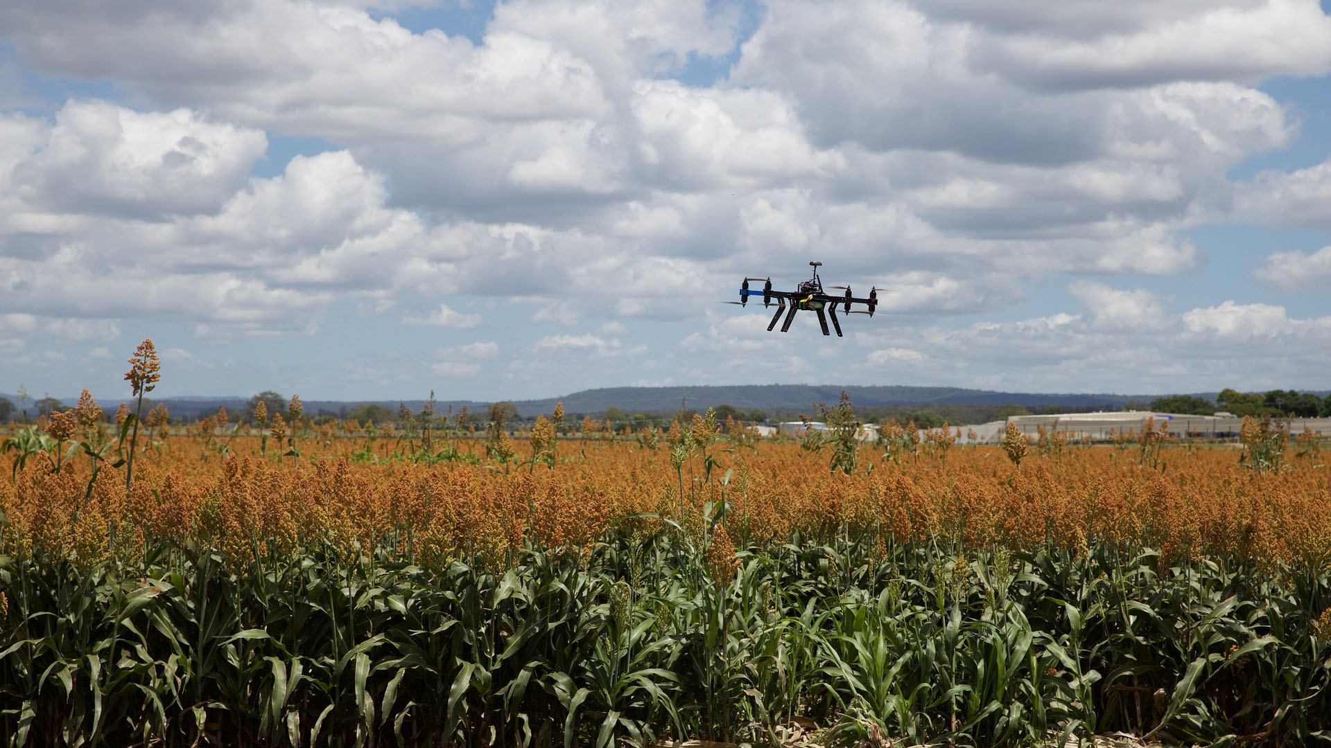A consumer drone hovering over a field against a sunny, but cloudy sky.