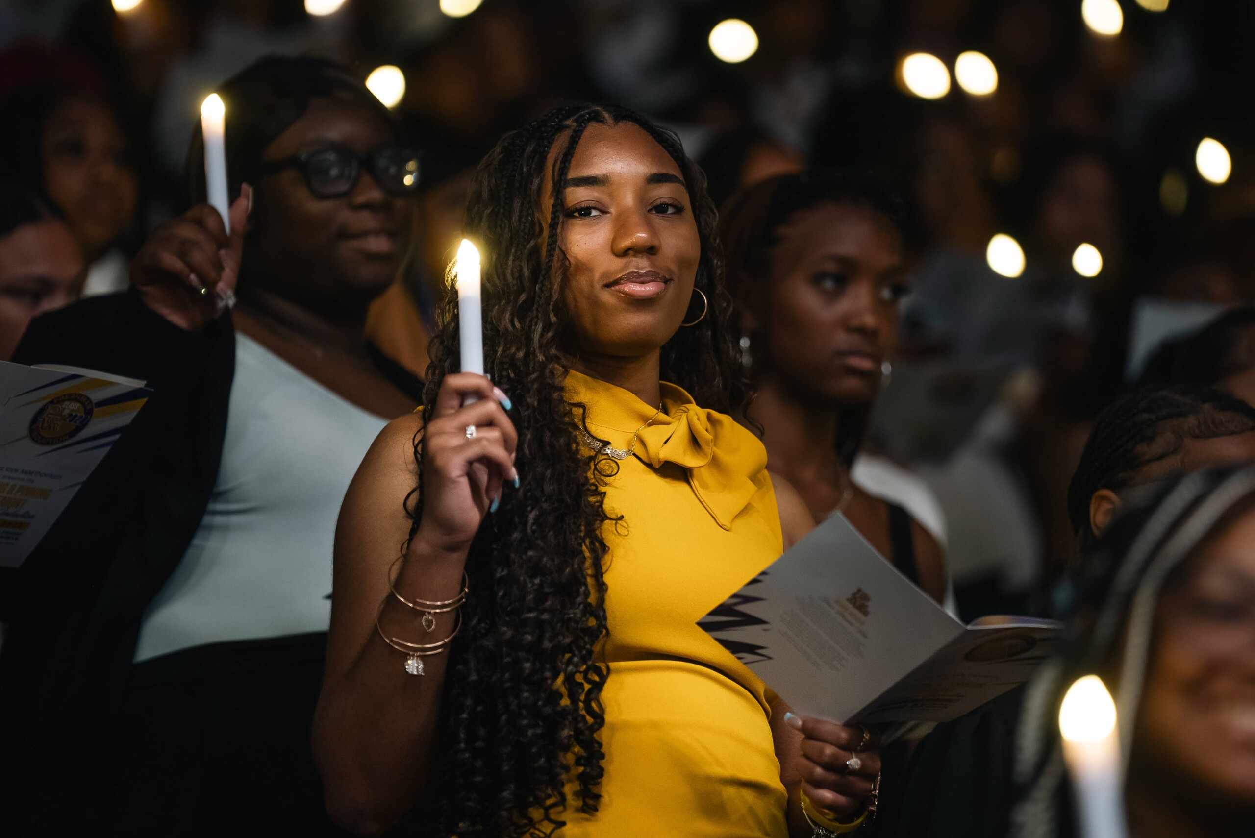 A woman in a yellow dress holds a candle and a program, surrounded by a crowd of similarly dressed individuals in a dimly lit setting.