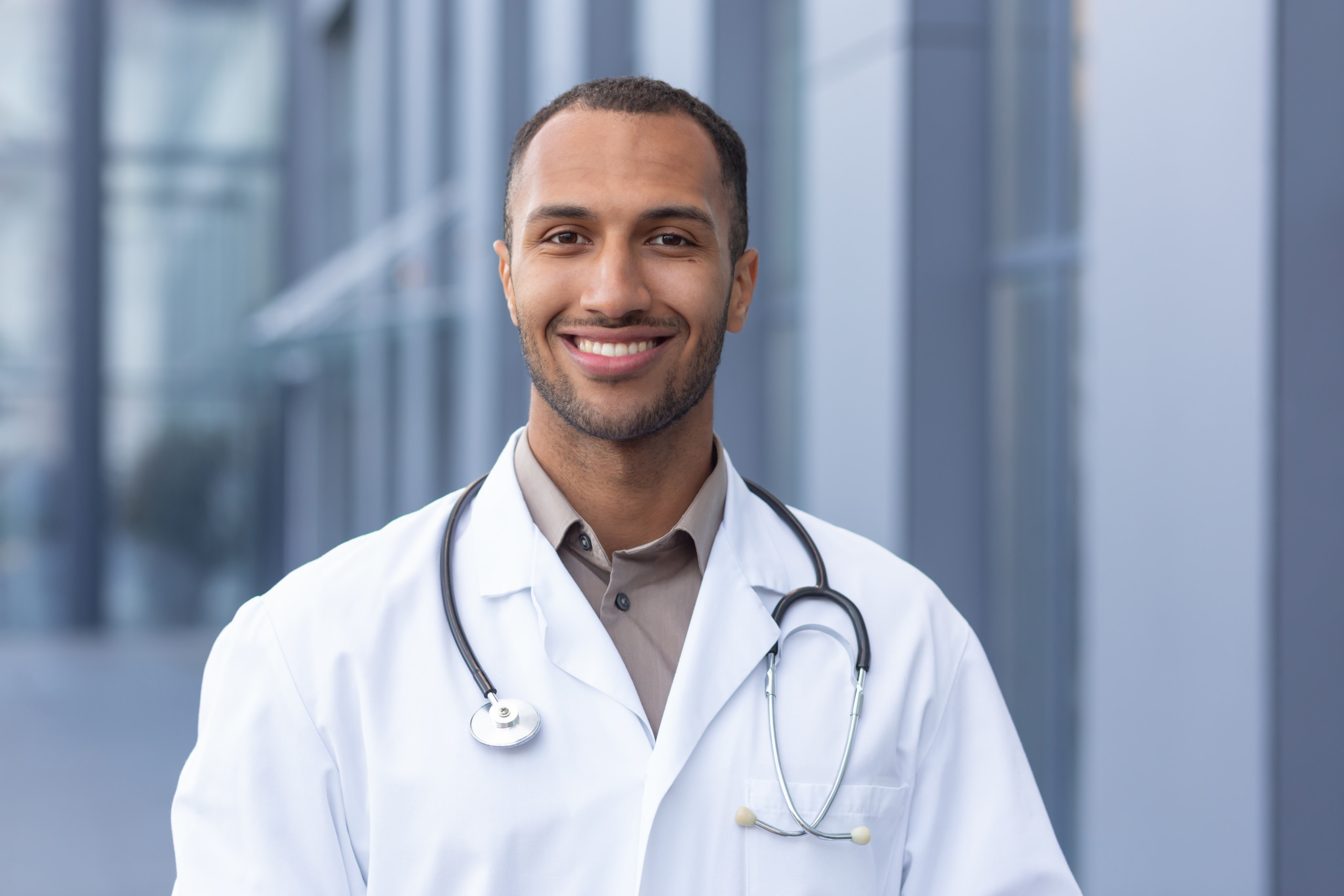 Portrait Of Successful And Smiling African American Doctor, Man In Medical Coat With Stethoscope Looking At Camera And Smiling, Outside Modern Hospital