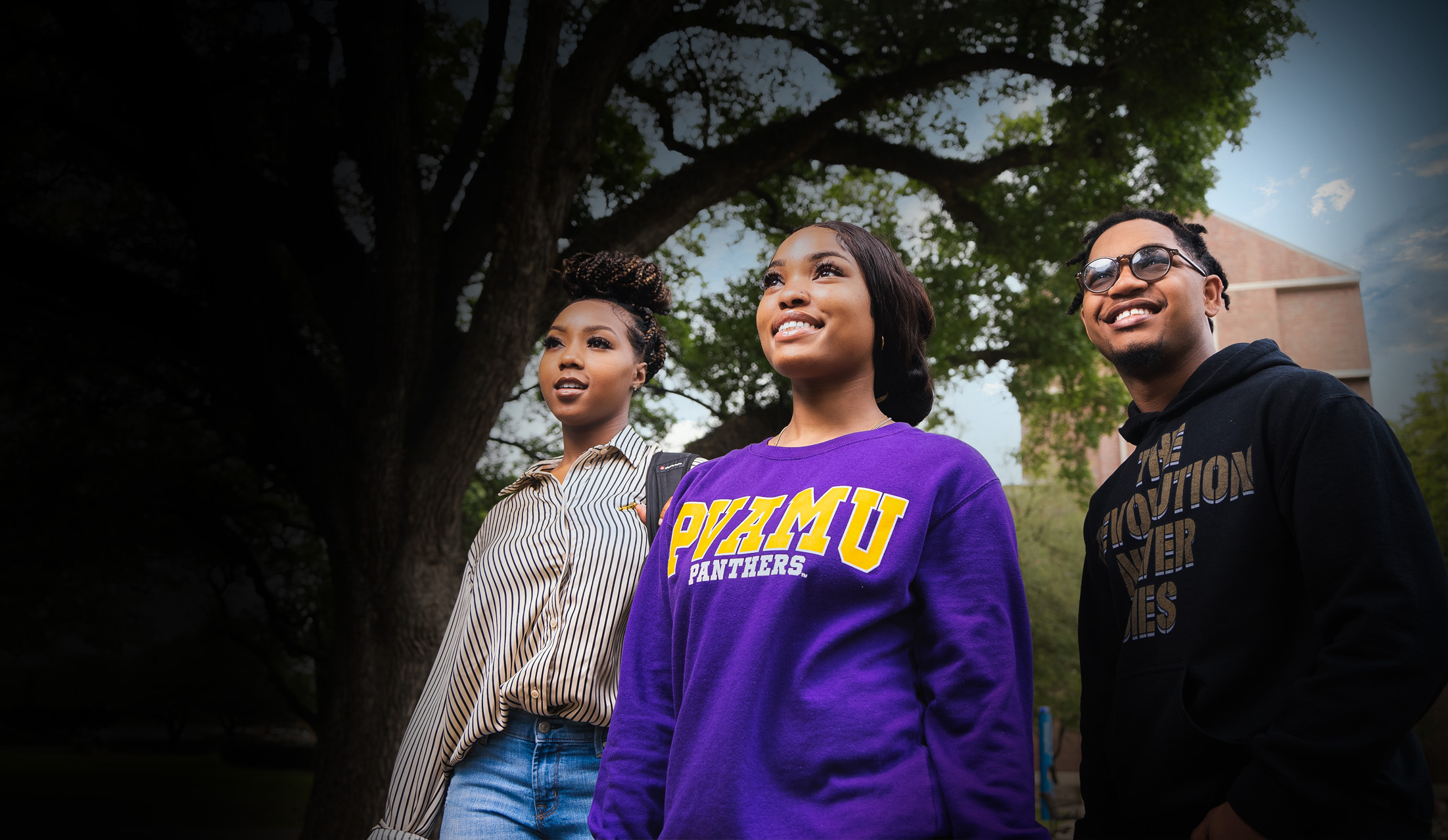 Admissions 4 Three students stand together outdoors, wearing casual clothing, with a large tree and a building visible in the background.