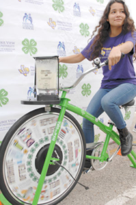 The newspaper feature shows Projecto Juan Diego Heroes-4-Health member Desiree Garcia making an apple smoothie while cycling during Proyecto Juan Diego World Diabetes Day Health Fair