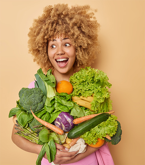 Woman with light brown curly hair smiling with joy while holding an overflowing armful of vegetables.