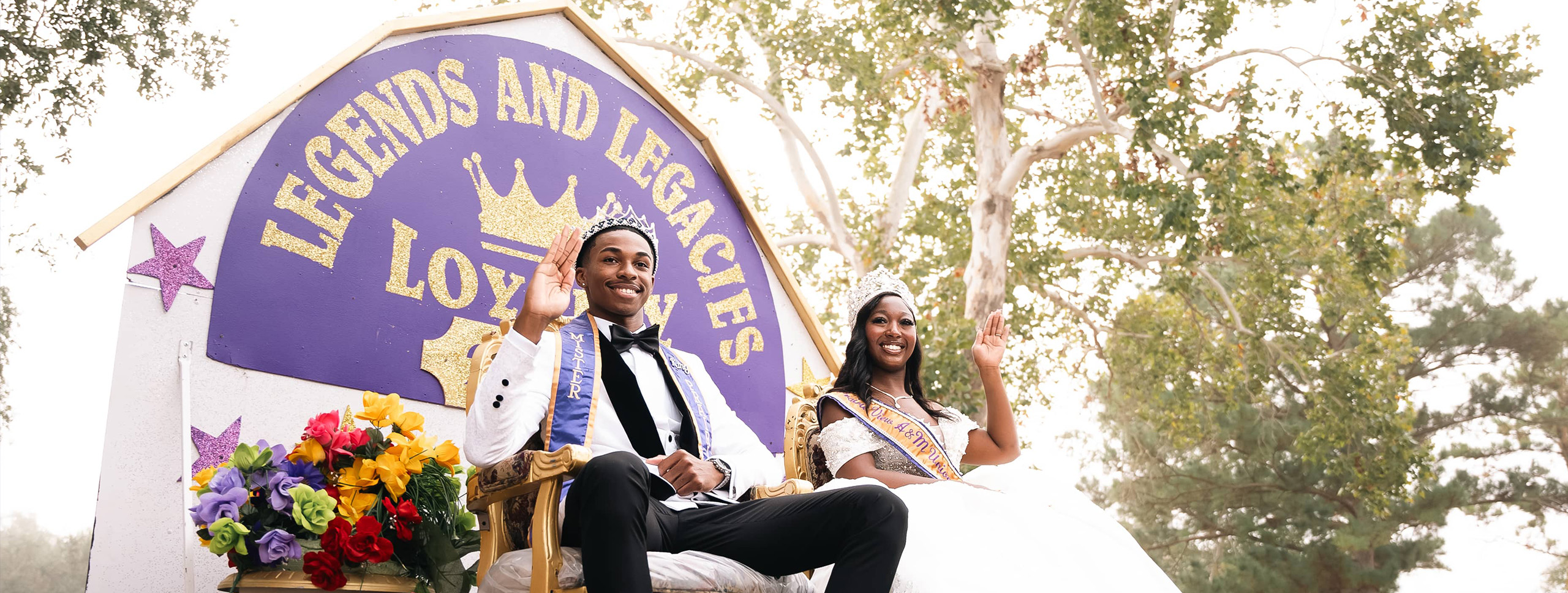 A crowned couple sits on a decorative throne, surrounded by flowers, waving to an audience at an outdoor event under trees.
