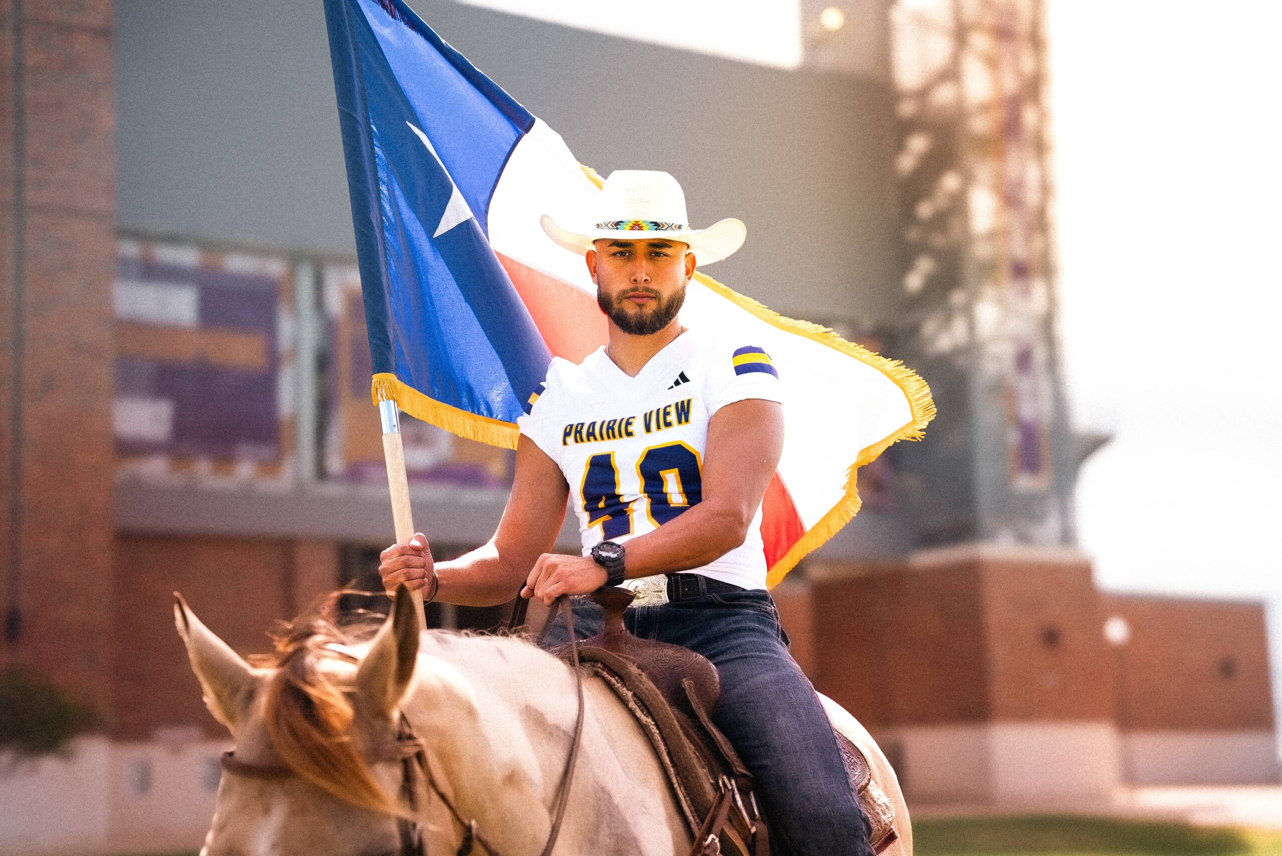 A football player in a white jersey holds a Texas flag, wearing a cowboy hat, with a vibrant backdrop.