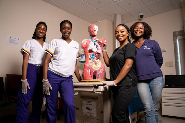 DSC01089 Four women in scrubs stand beside a detailed anatomical model in a clinical setting, showcasing their involvement in healthcare education.