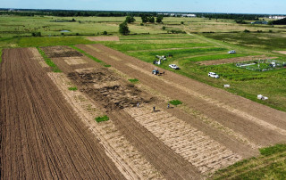 Aerial view of agricultural research plots with tilled fields, green pasture, farm vehicles, equipment and workers spread across a rural landscape.