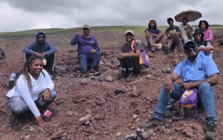 A group of people smiles while sitting on a rocky hill under cloudy skies. They appear relaxed and joyful, suggesting a friendly, adventurous outing.
