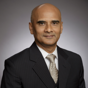 Professional headshot of a man in a dark suit, white shirt and patterned tie against a neutral gray background.