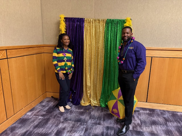 2 staff members standing in front of Mardi Gras themed backdrop