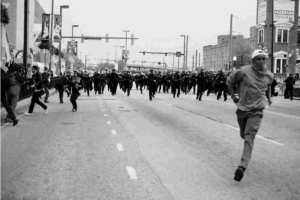 Riot in Baltimore, man running from a line of charging police.